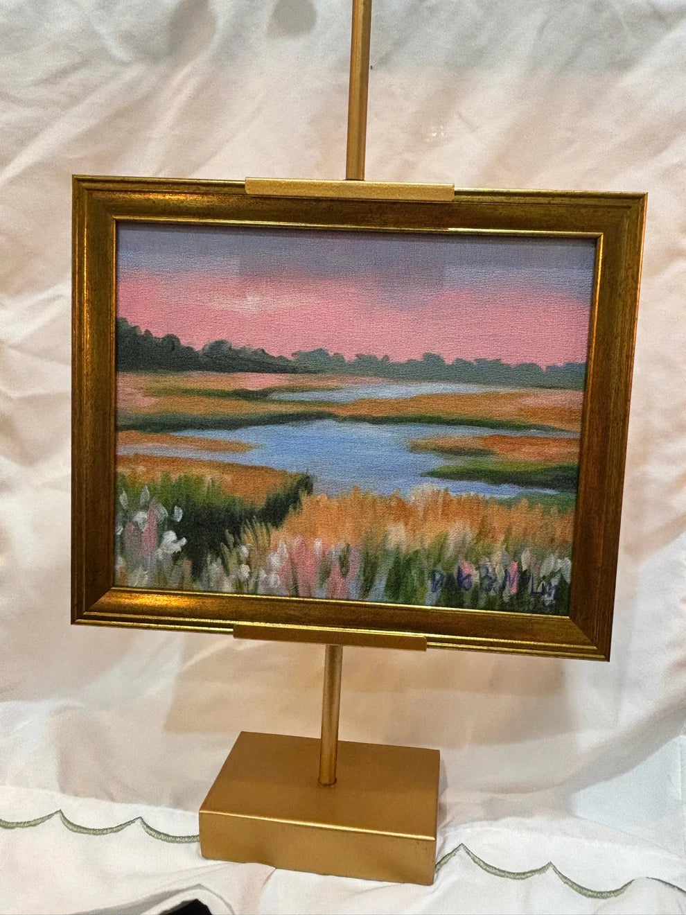 Painting of a landscape with a lake and fields on a gold easel against a white background