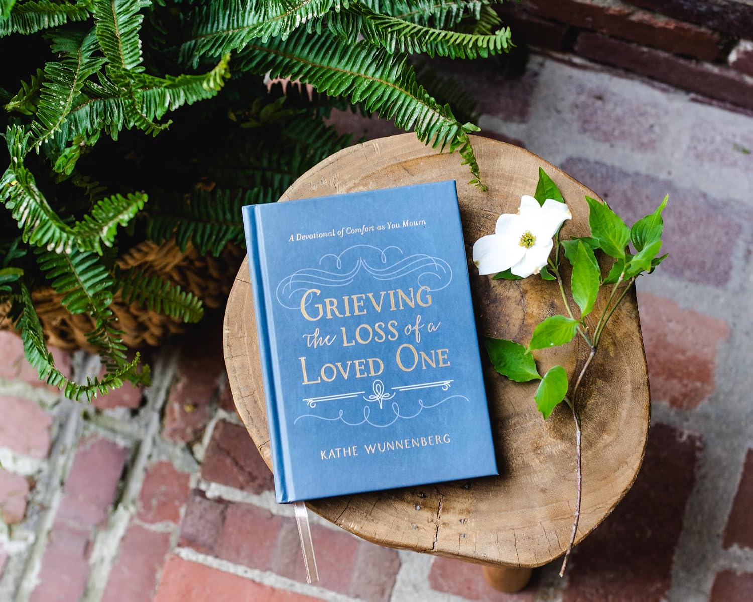 Blue book titled 'Grieving the Loss of a Loved One' by Kathie Wunnenberg on a wooden stool with greenery.