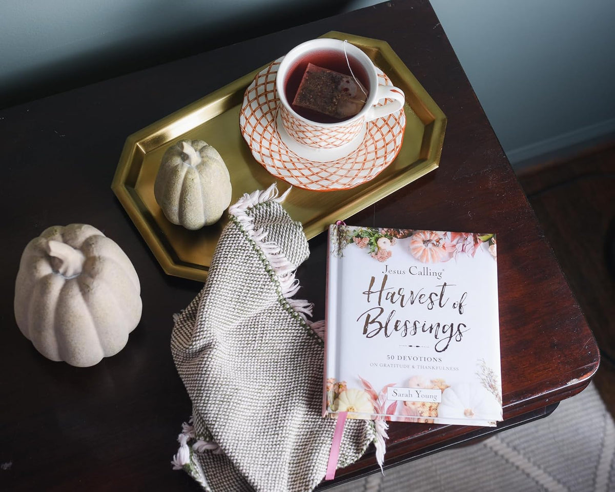Teacup with a tea bag, decorative pumpkins, and a book titled 'Harvest of Blessings' on a wooden table.