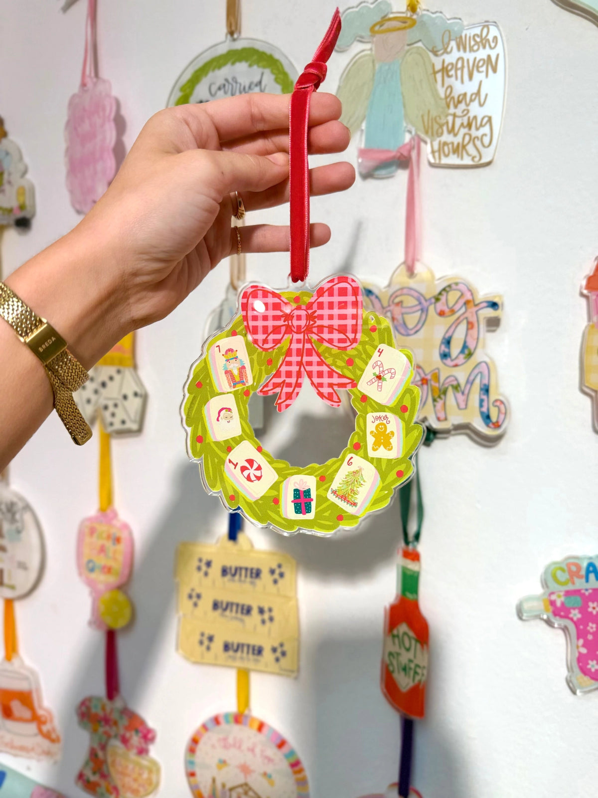 Hand holding a colorful wreath-shaped ornament with a gold watch on wrist, against a wall of similar ornaments.