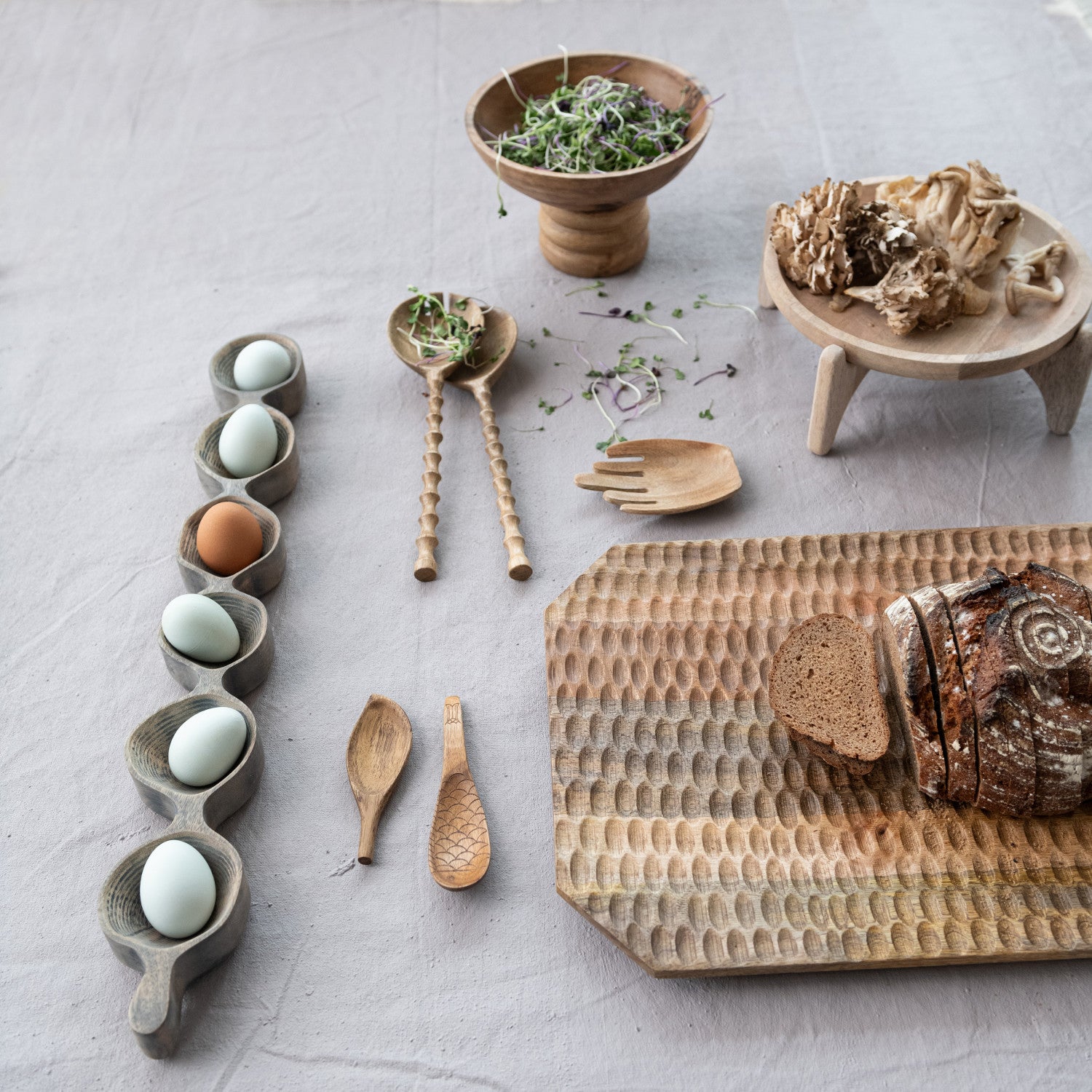 Wooden egg holder with eggs, wooden spoons, and a loaf of bread on a light gray surface.