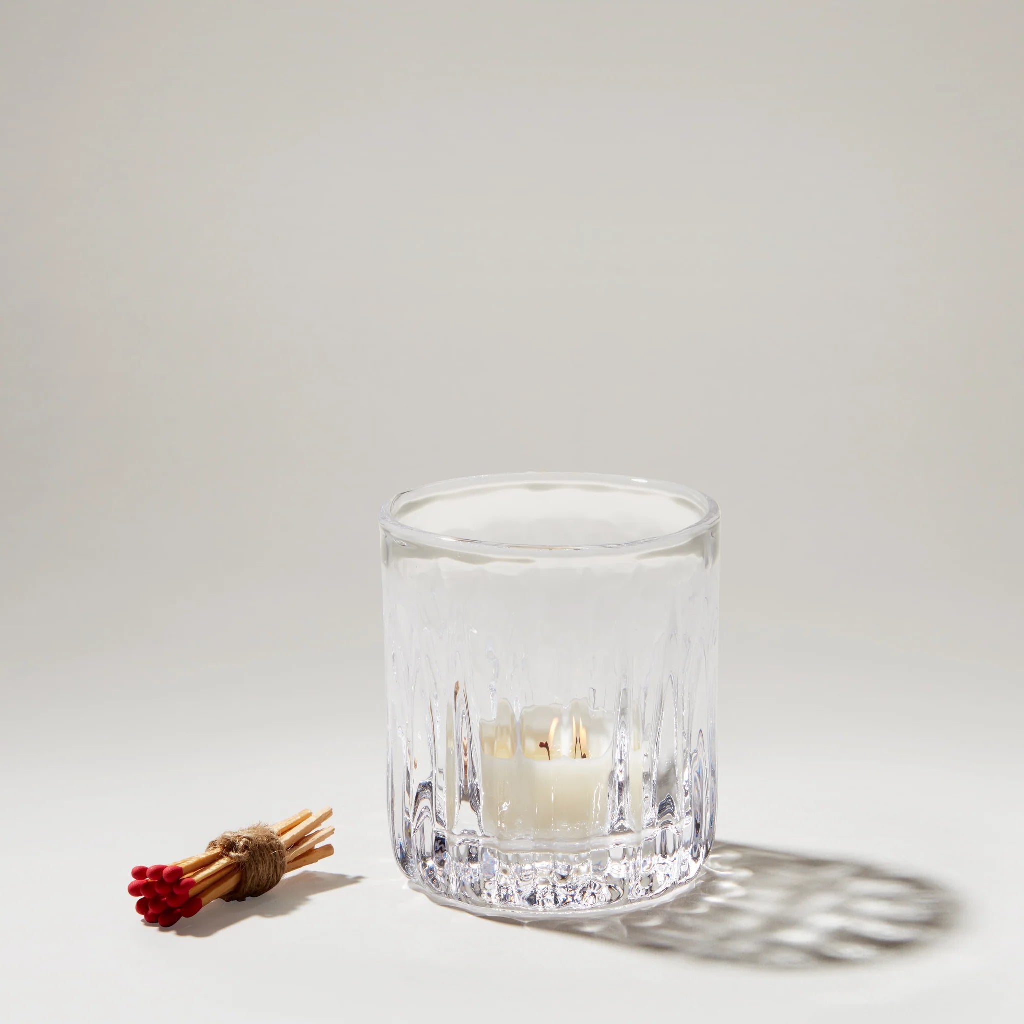 Clear glass candle holder with a lit candle and matches on a light gray background