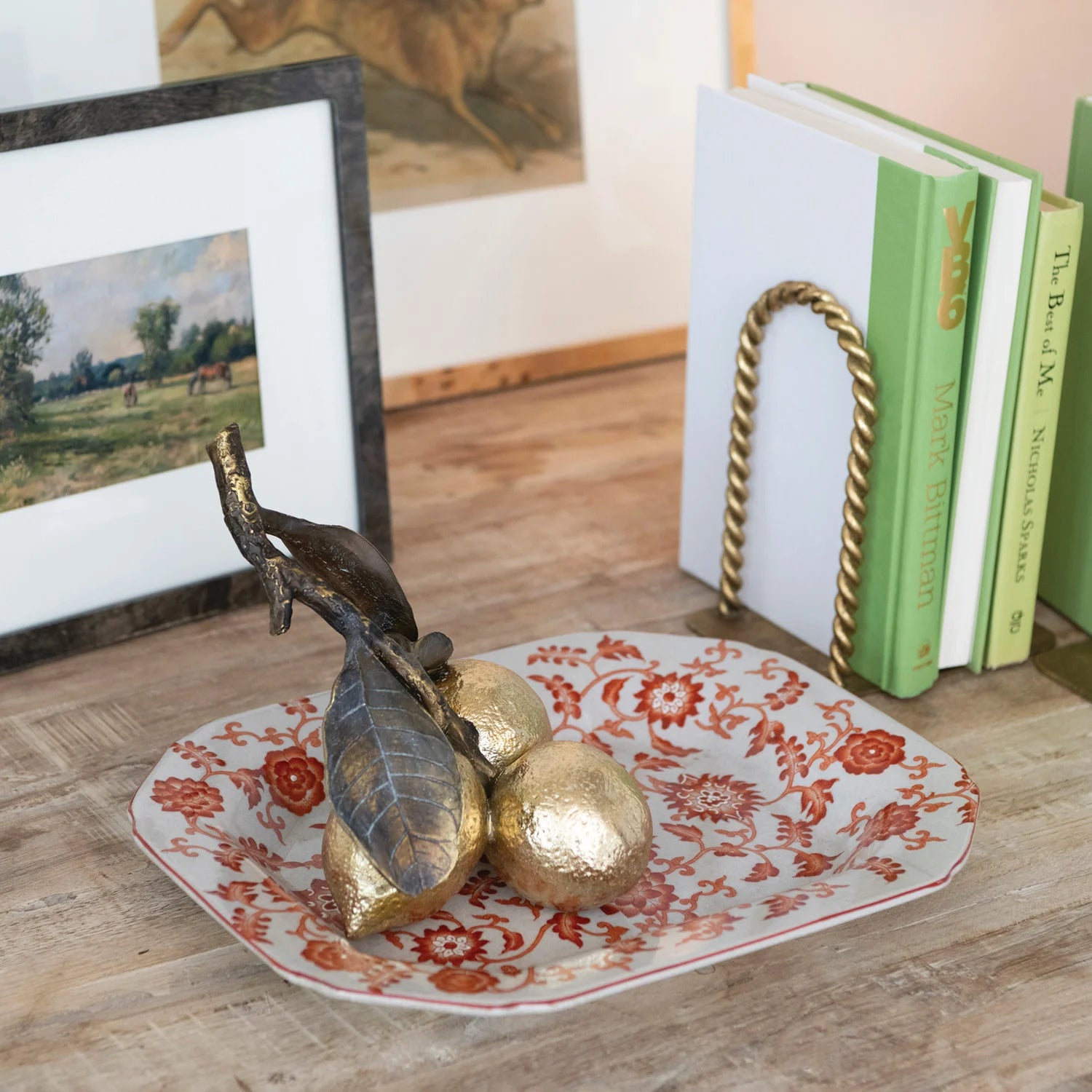 Decorative plate with gold leaves and a bird on a wooden surface with books and a painting in the background.