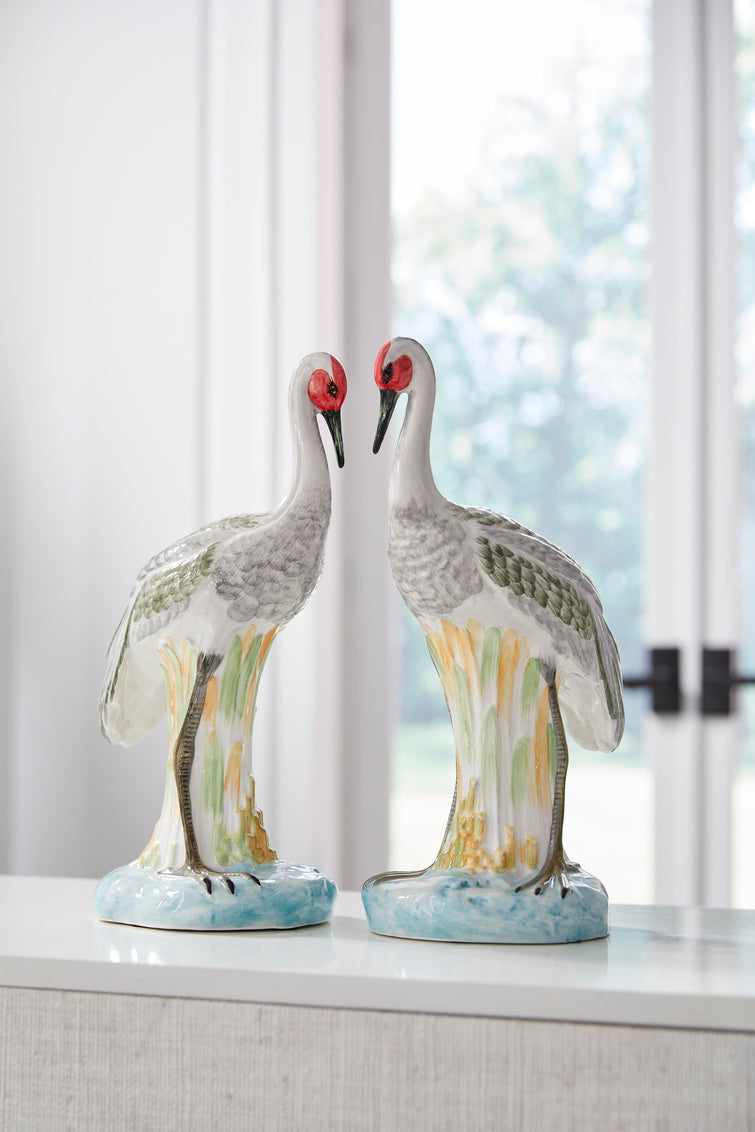 Two ceramic crane figurines on a surface with a blurred window background
