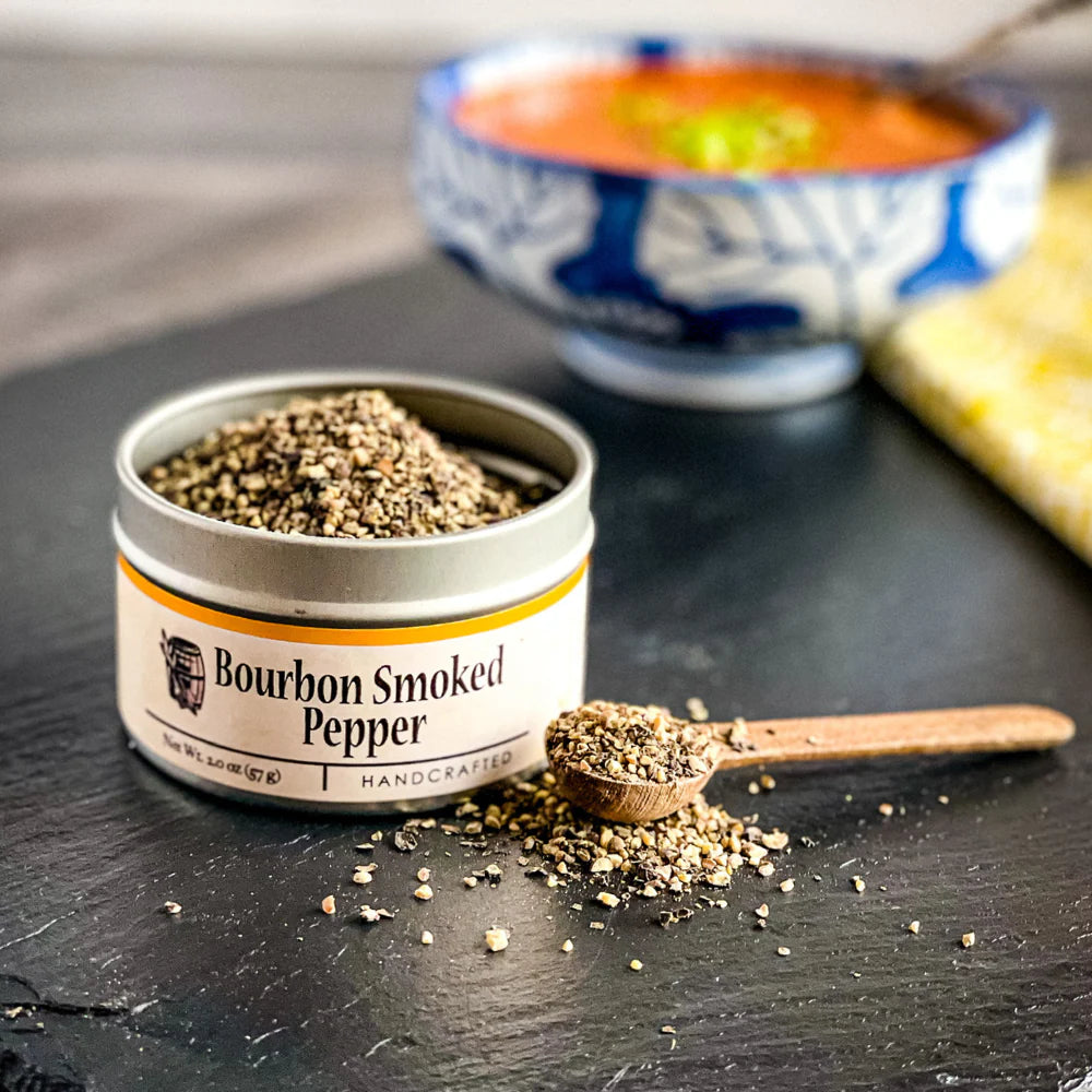 Bourbon Smoked Pepper container with a wooden spoon on a dark surface, with a blurred bowl of soup in the background.