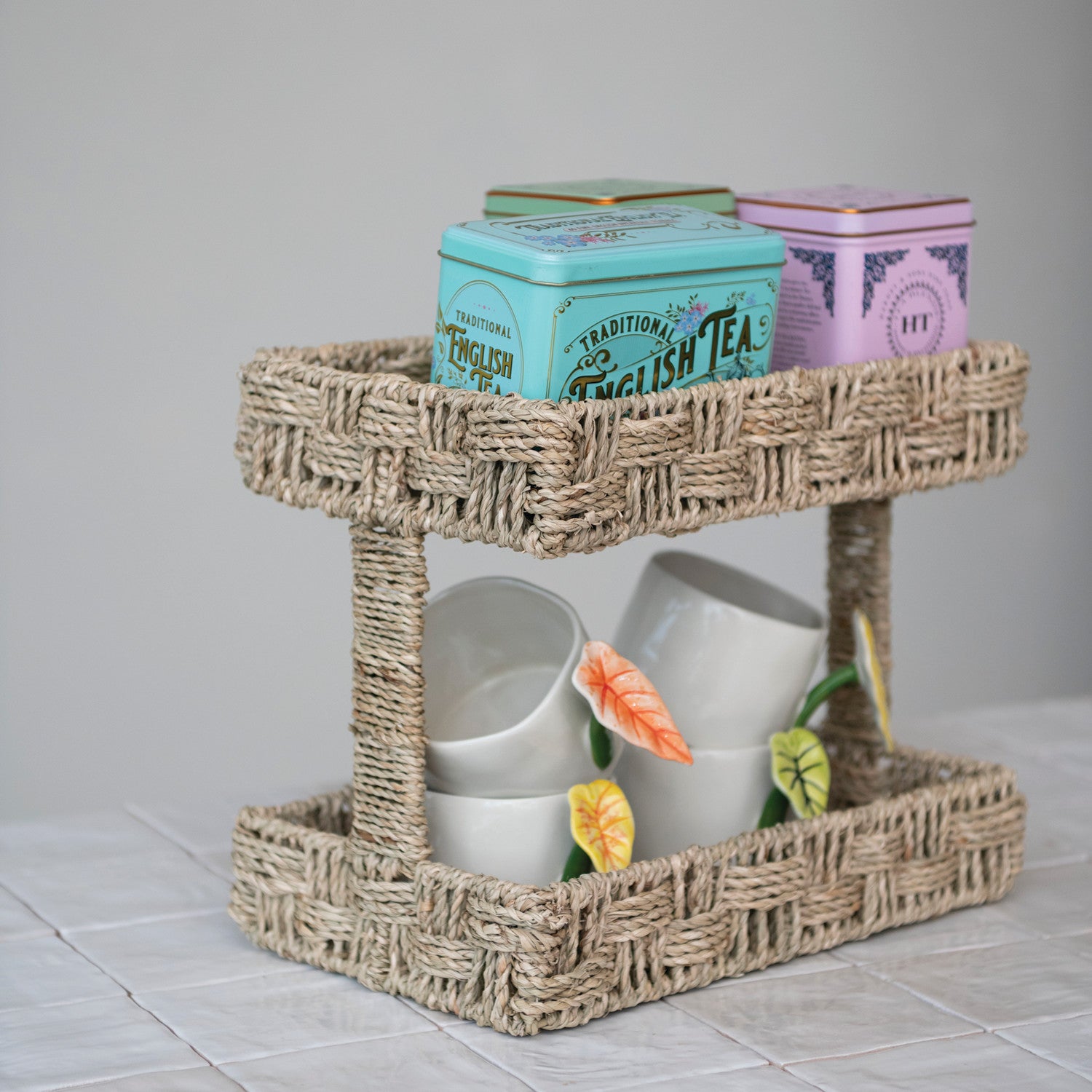 Woven two-tier tray with tea cups and tins on a tiled surface