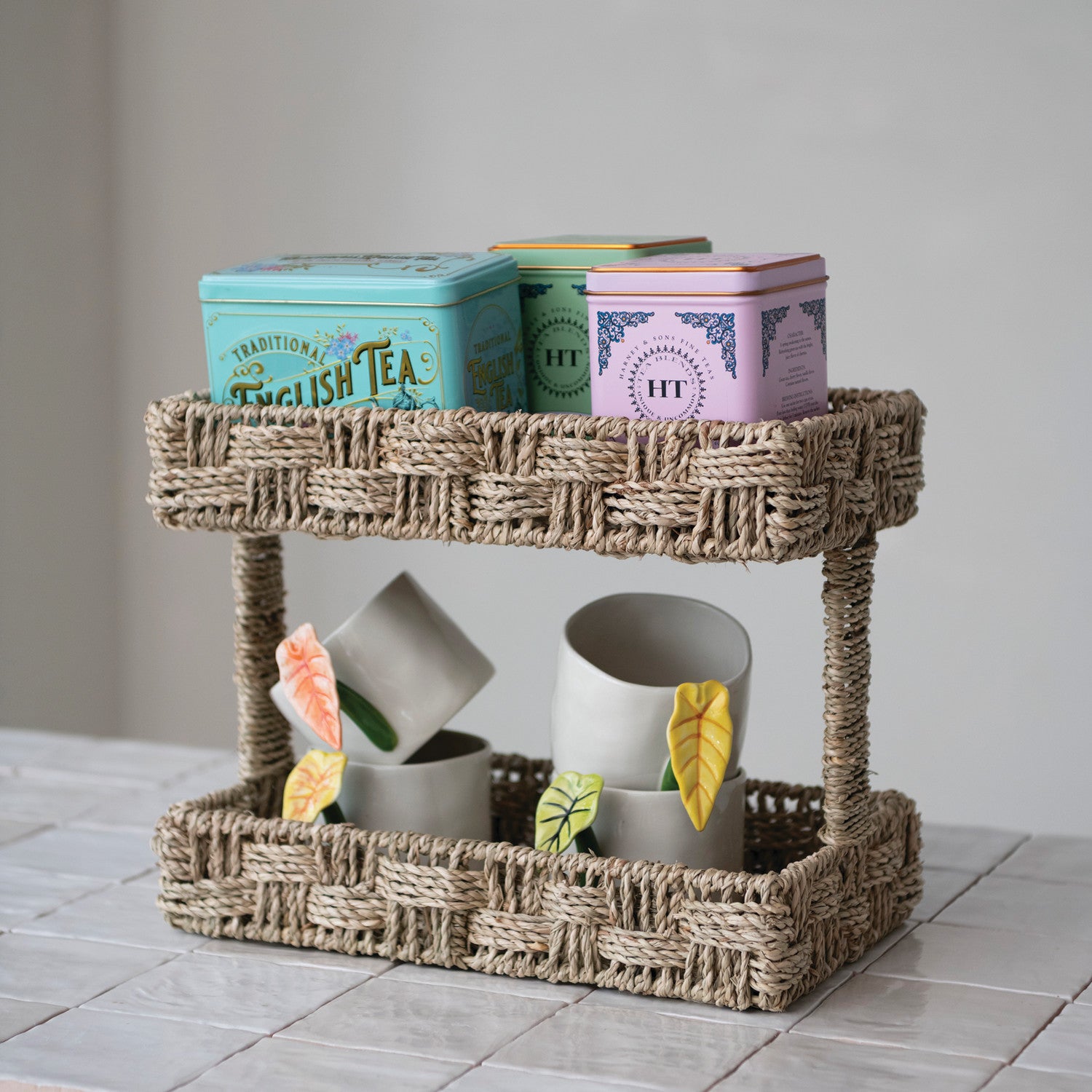 Two-tier woven tray with tea tins and mugs on a tiled surface.