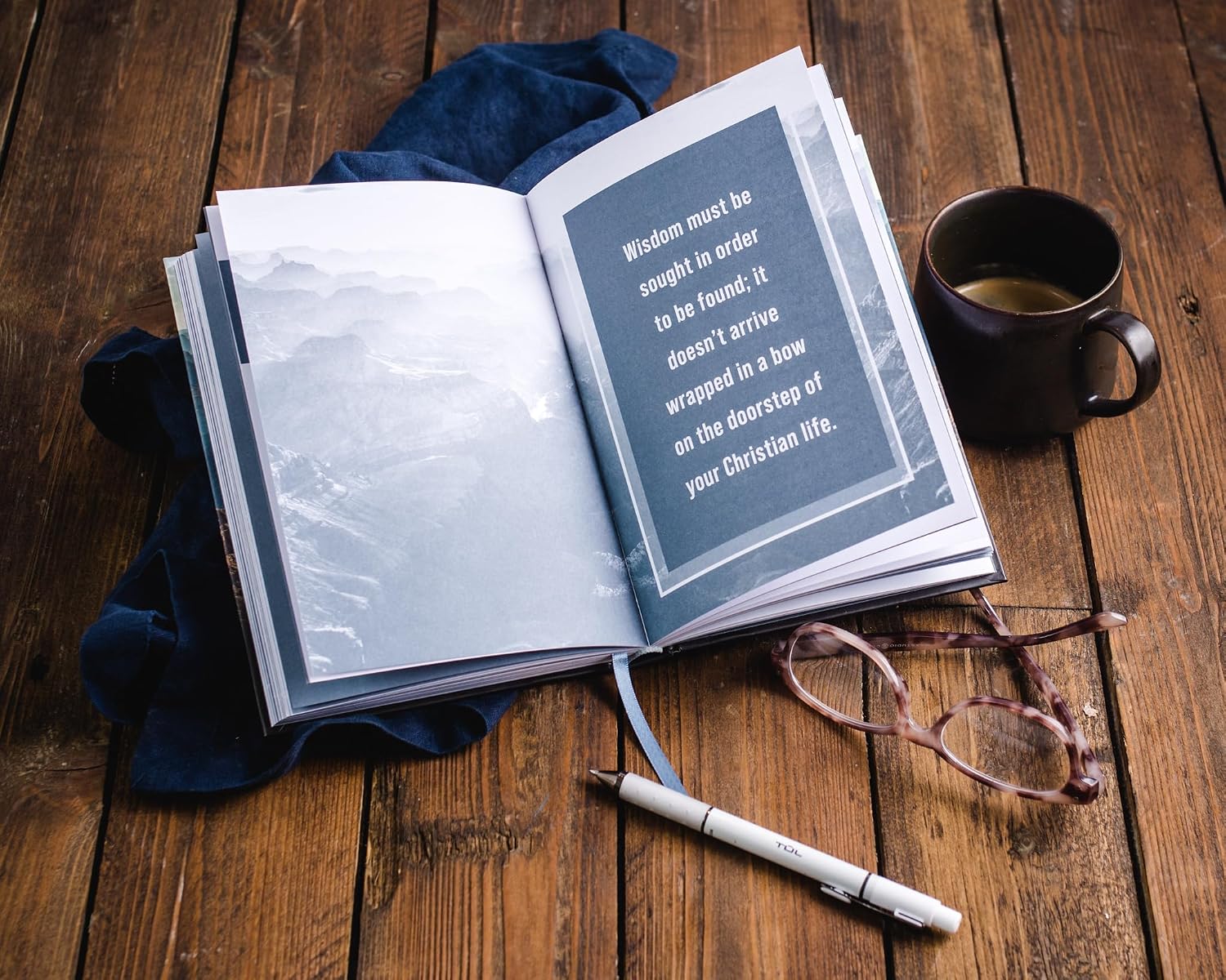 Open book with a quote on a wooden surface, accompanied by a mug and glasses.