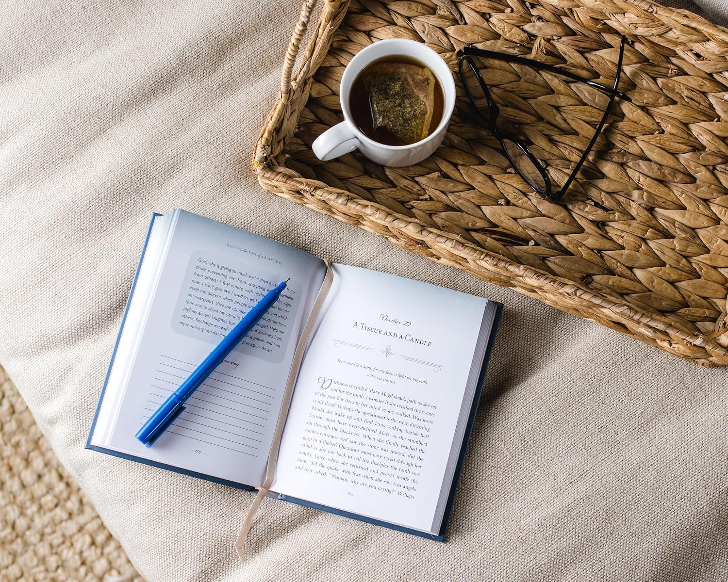Open book with a pen, mug of tea, and glasses on a woven tray.