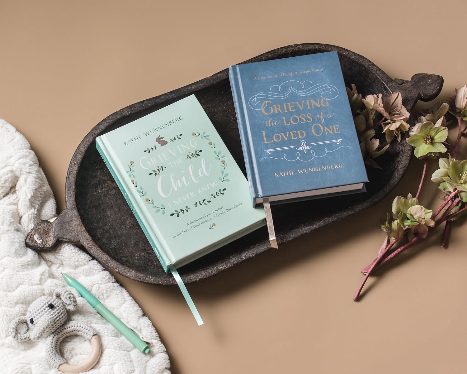 Two books titled 'My Child' and 'Grieving the Loss of a Loved One' on a dark wooden tray with flowers and a baby rattle.