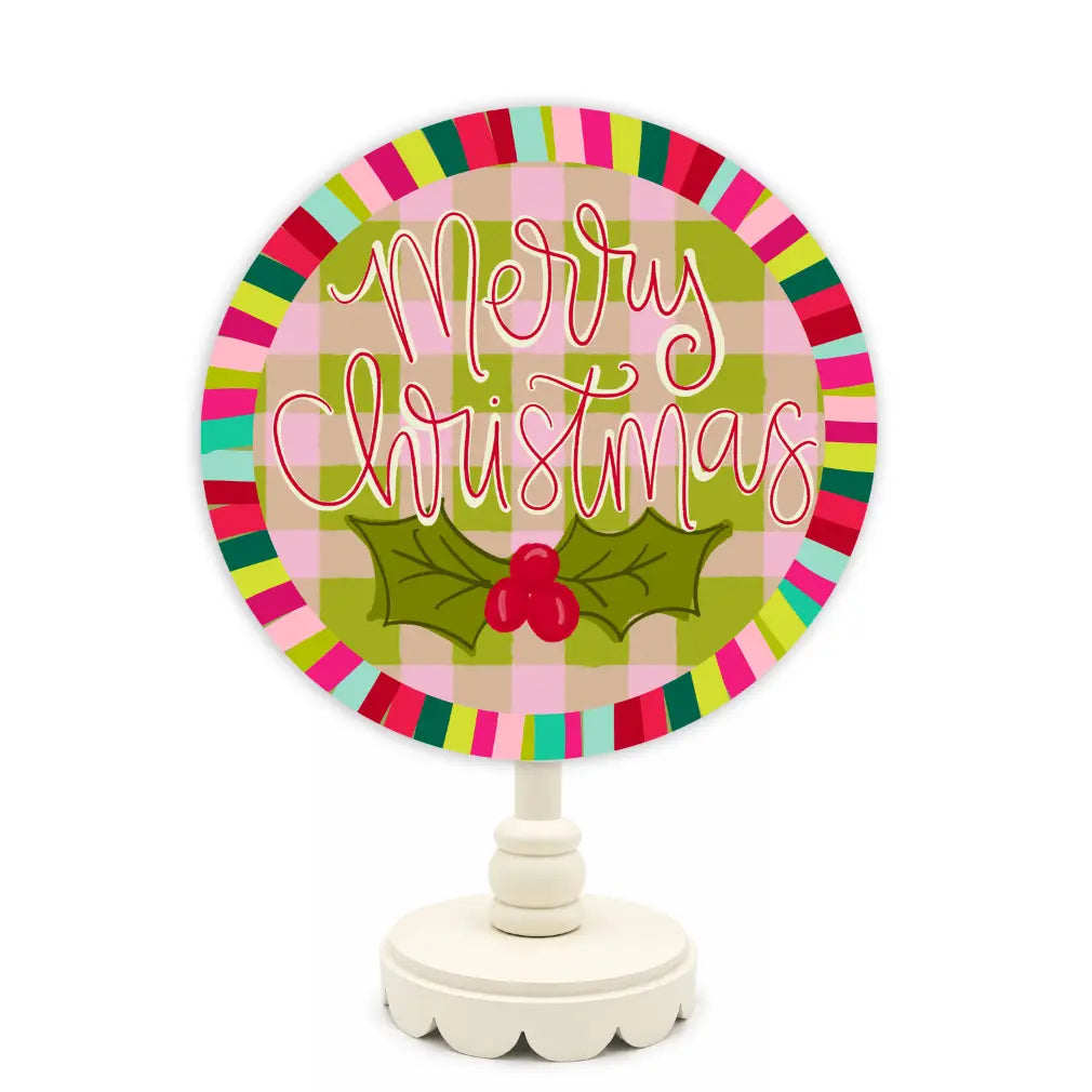 A round tabletop Christmas sign on a white stand, featuring a pink and green plaid background, a colorful striped border, and the words 'Merry Christmas' in white script above a green holly leaf with red berries.