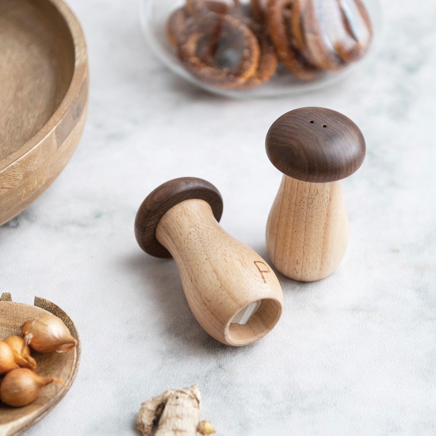 Wooden mushroom-shaped salt and pepper shakers on a marble surface with ingredients in the background.