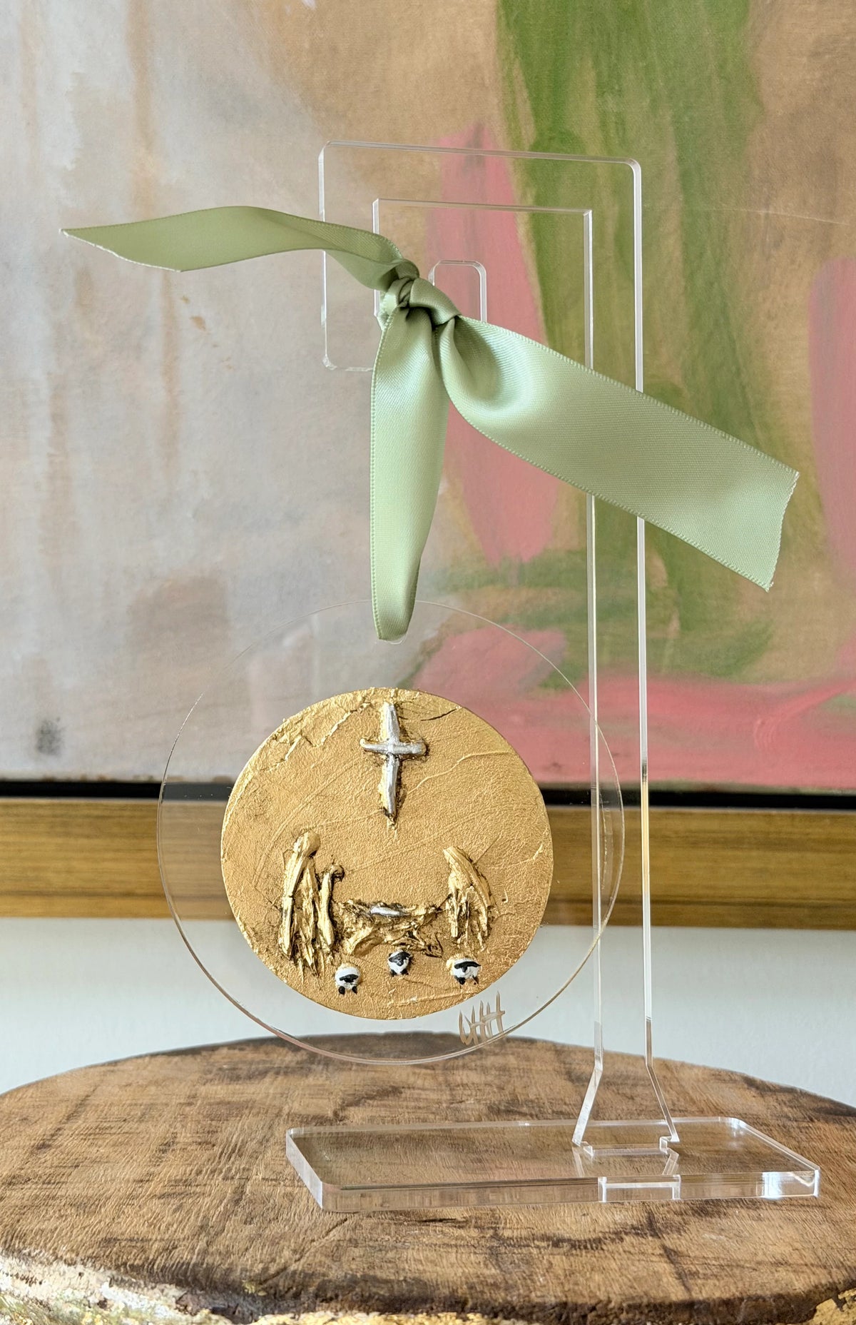 A round gold-toned decorative plaque or medallion with embossed religious figures and a cross, displayed on a clear acrylic stand with a pale green ribbon bow. The stand sits on a rustic wood slice in front of a softly blurred abstract painting.