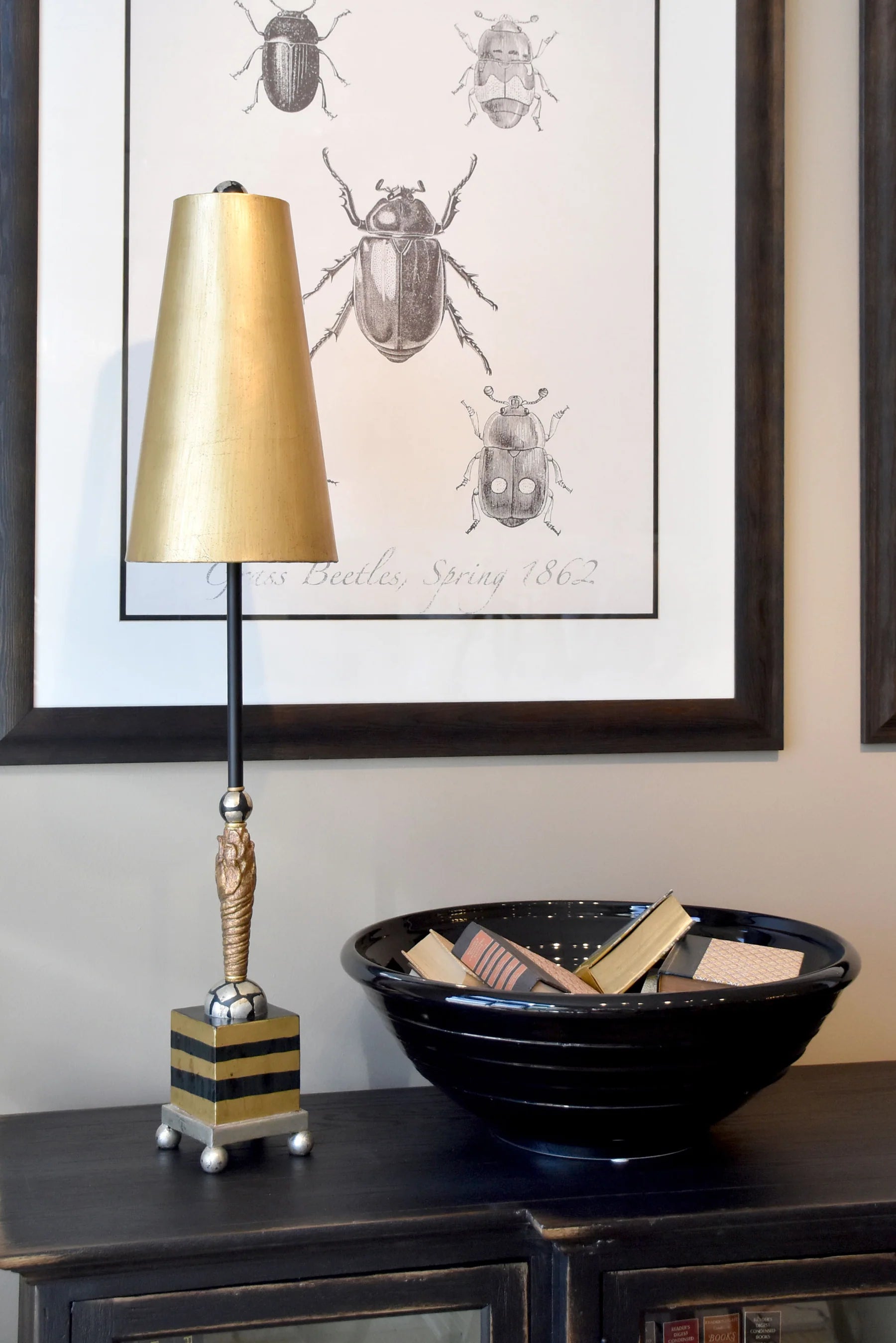 Decorative lamp with gold shade and striped base on a shelf with a black bowl and framed beetle art in the background.